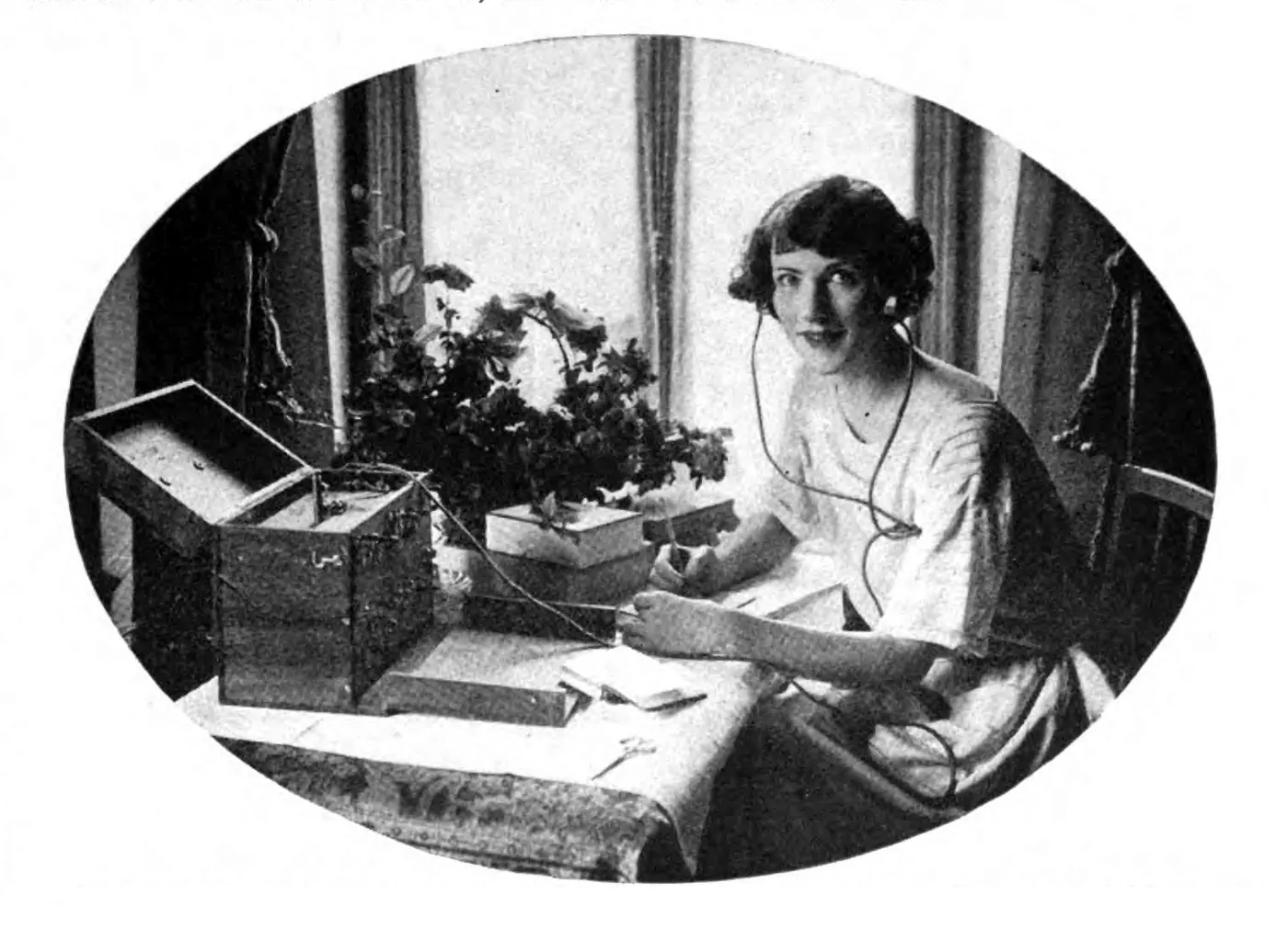 A young woman at a desk wearing a headset wired to a small radio receiver, 1923